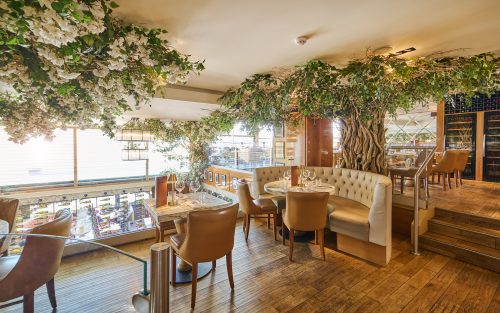 Upscale restaurant dining room with hanging green vines and white flowers overhead, round marble tables and beige upholstered chairs.