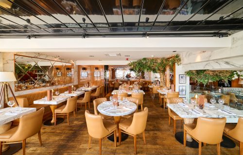 Elegant restaurant dining room with marble-topped tables, tan leather chairs, and warm ambient lighting across wooden floors.