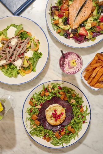 A colorful assortment of salads on blue-rimmed plates: beef with greens and cheese, a salmon and fruit salad, and a beet carpaccio with arugula and pomegranate seeds.