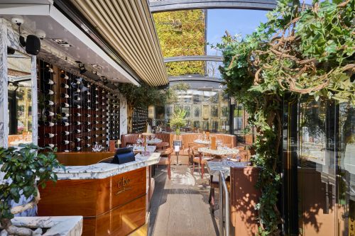 Upscale restaurant interior with a wine-wall display, marble counter, tufted leather booths, and greenery framing glass walls.