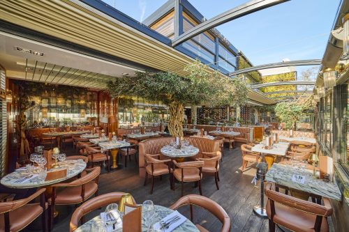 Upscale restaurant dining area with marble-topped tables, brown leather chairs, and indoor trees under glass skylights.