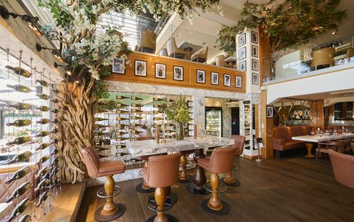 Upscale wine bar with a large twisted tree centerpiece, marble counters, and wall-mounted wine racks; leather stools and framed photos along a wood-paneled wall.