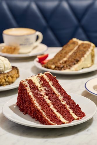Slice of red velvet cake with cream cheese frosting on a white plate in the foreground; a coffee cup and more cake slices are blurred in the background on a table.