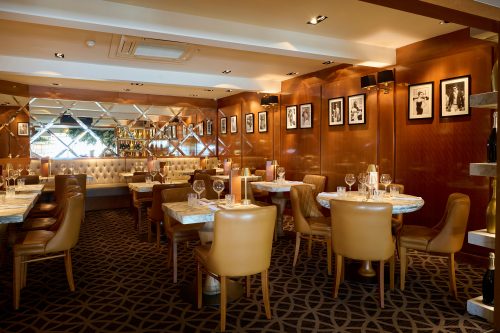 Elegant restaurant dining area with marble-topped tables, tan leather chairs, wine glasses, and framed black-and-white photos on wood-paneled walls (informative).