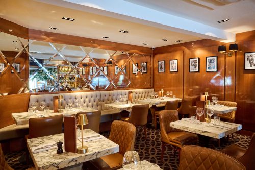 Upscale restaurant interior with marble tables, brown leather chairs, and mirrored lattice dividers along wood-paneled walls with framed photos