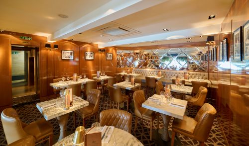 Elegant restaurant dining room with marble-top tables, tan leather chairs, and warm lighting reflecting off mirrored walls.