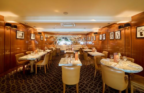 Elegant dining room with marble tables, brown leather chairs, framed photos on wood-paneled walls, and warm lighting
