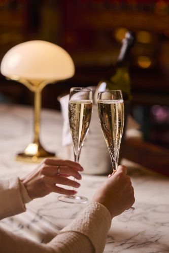 Two people clinking champagne flutes on a marble table with a blurred bottle and lamp in the background.