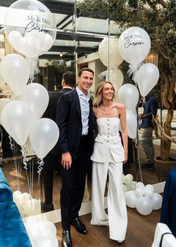 Smiling couple in formal attire posing among white balloons at a wedding venue, with balloons reading 'Mr & Mrs Carson'