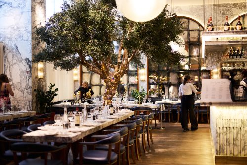 Elegant restaurant interior featuring a large illuminated tree centerpiece, marble walls, and formally set dining tables with wine glasses.