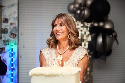 Smiling woman at a birthday party with a frosted cake in front and balloon decorations in the background.
