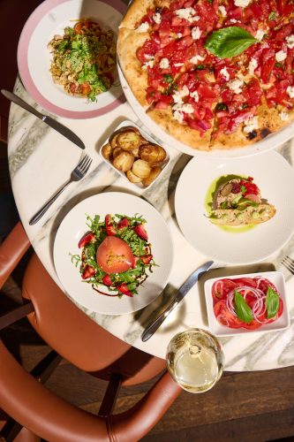 Overhead view of a dining table with a large pizza topped with tomatoes and cheese, plus sides like salads and roasted potatoes, and a glass of white wine.
