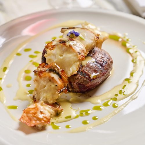 Lobster tail on a seared steak with creamy sauce and a purple flower garnish on a white plate, with green oil accents around the rim