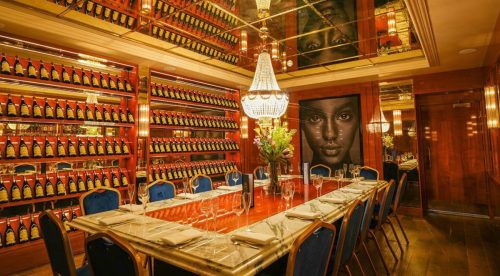 Luxurious private dining room with champagne bottles lining red shelves, a crystal chandelier, and a long marble table set for a meal.