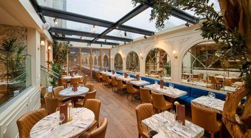 Bright, elegant restaurant interior with marble-top tables, tan quilted chairs, and a glass ceiling corridor lined with plants and arches.