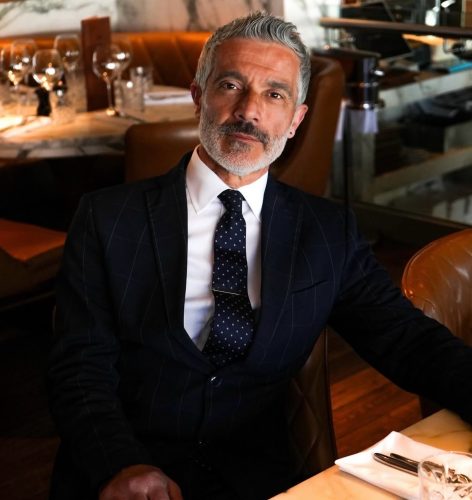 Man in a dark pinstripe suit and navy polka dot tie sits at a marble table in a restaurant, looking at the camera with a smile-yet-serious expression.