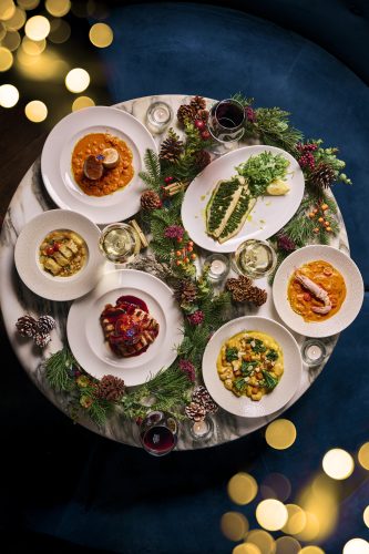 Overhead view of a festive table with several plated dishes, wine glasses, greenery and pinecones under warm bokeh lights.