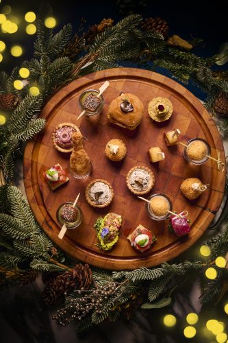 Assorted bite-sized appetizers arranged on a round wooden board among evergreen branches and pinecones.