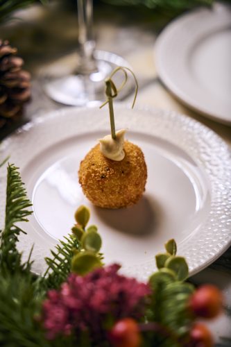 Golden fried croquette on a white plate, topped with a dollop of creamy sauce and skewered with a decorative pick on a festive table setting.
