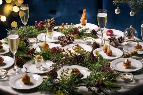 Festive holiday dinner table with champagne flutes, white plates, canapés, pinecones, and greenery.