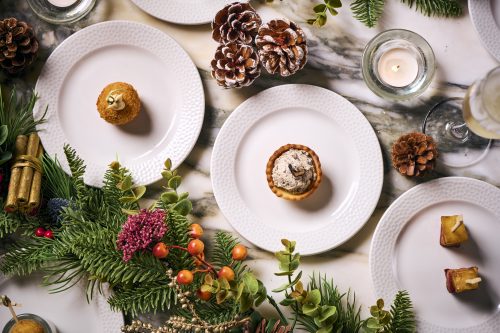 Top-down view of a festive table setting with white textured plates, pastries, pinecones, candles, and evergreen garlands.