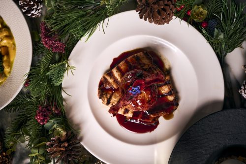 Grilled meat slices with a red glaze on a white plate, surrounded by festive evergreen garlands and pinecones.
