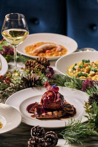 Festive table setting with a glazed roast on a white plate, sauce pooling around, and a glass of white wine nearby amid pine cones and greenery.