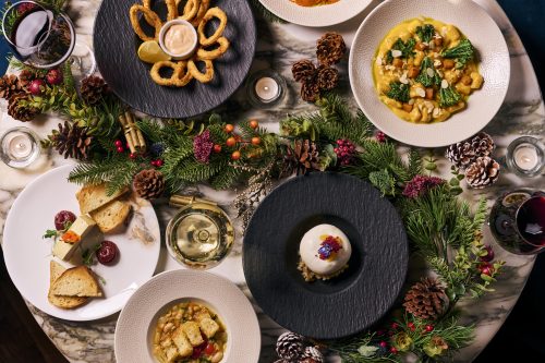 Top-down view of a festive holiday table with bowls of soup, bread, and appetizers surrounded by evergreen garlands, pine cones, and candles on a marble table.