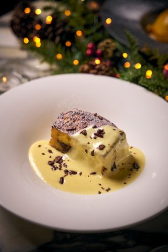 Close-up of a sliced cake on a white plate, covered in creamy sauce and chocolate shavings, with festive bokeh lights in the background
