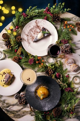 Top-down view of a festive table with a slice of chocolate cake on a white plate, cocoa dusting, and holiday greenery with pine cones and berries