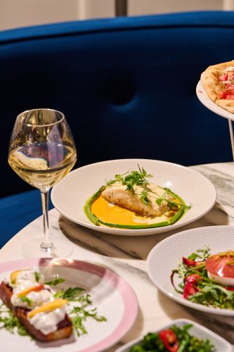 Fish fillet in orange sauce with green herb garnish on a white plate; a glass of white wine nearby with other dishes on a marble table.