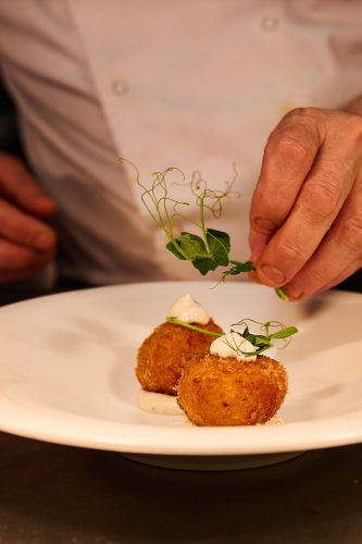 Chef’s hands garnish two crispy croquettes with dill and dollops of cream on a white plate.