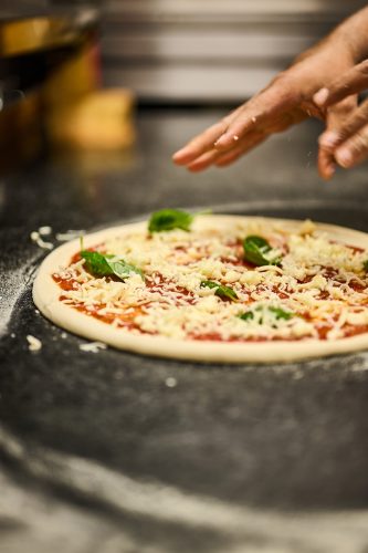 Pizza ready to bake with tomato sauce, mozzarella, and basil leaves, while someone sprinkles cheese on top.