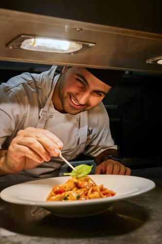 Chef in a white coat and black toque carefully garnishing a plated pasta dish under kitchen lighting.