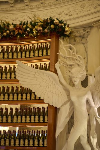 White marble angel sculpture with large wings beside rows of wine bottles and festive garland on an ornate wall interior.