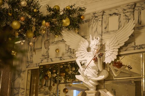 White angel sculpture playing a violin beneath gold Christmas ornaments and evergreen branches on a marble wall backdrop.