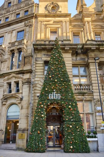 Tall Christmas tree decorated with gold and red baubles in front of an ornate sandstone building with arched windows and balconies.