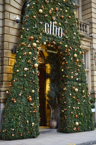 Decorated evergreen arch around a restaurant entrance with gold, bronze, and red ornaments and warm lights.