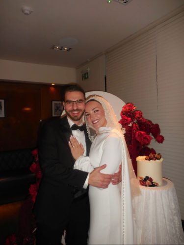 Bride in a white gown and veil hugs a man in a black tux at a wedding reception, with a tiered cake and red floral decor nearby.