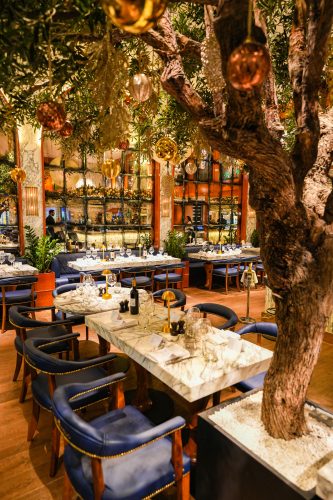 Elegant restaurant dining area with marble tables, blue upholstered chairs, and a large indoor tree adorned with gold ornaments.