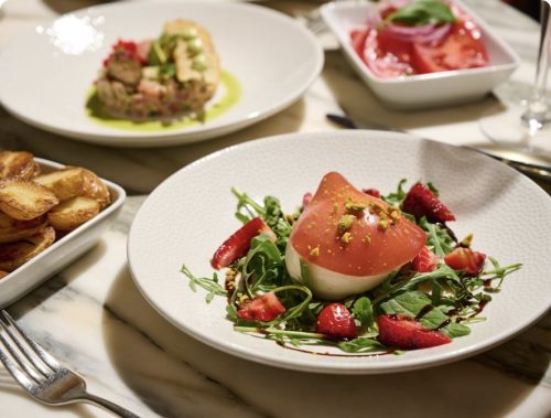 Plated salad with burrata topped by a tomato slice, arugula, strawberry pieces, pistachio crumbs, and balsamic drizzle. Side of roasted potatoes visible nearby in a white dish.