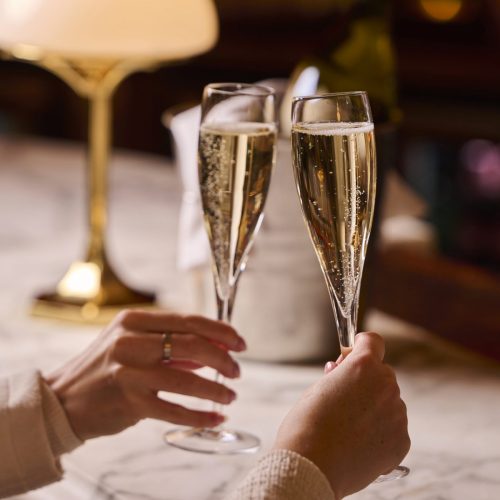 Two people toast with champagne flutes, hands visible and a warm, dimly lit setting in the background.