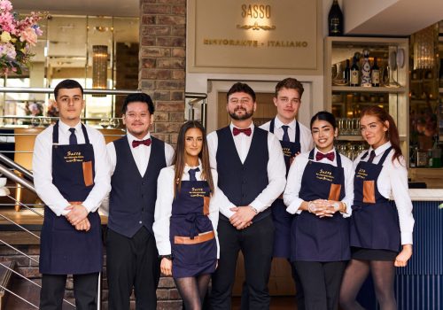 Group of seven restaurant staff in uniform, standing indoors in front of a sign that reads SASSO Ristorante Italiano.