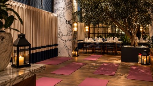 Elegant indoor space with pink yoga mats laid on a wooden floor, marble walls, and a large tree in a planter beside a candlelit dining area.